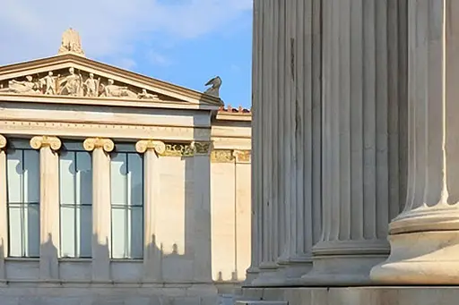 Sunlit classical building with columns and ornate carvings under a clear blue sky. card banner image
