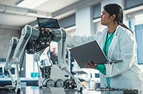A female robotics engineer in a lab coat using a laptop while examining a four-legged robot dog. 