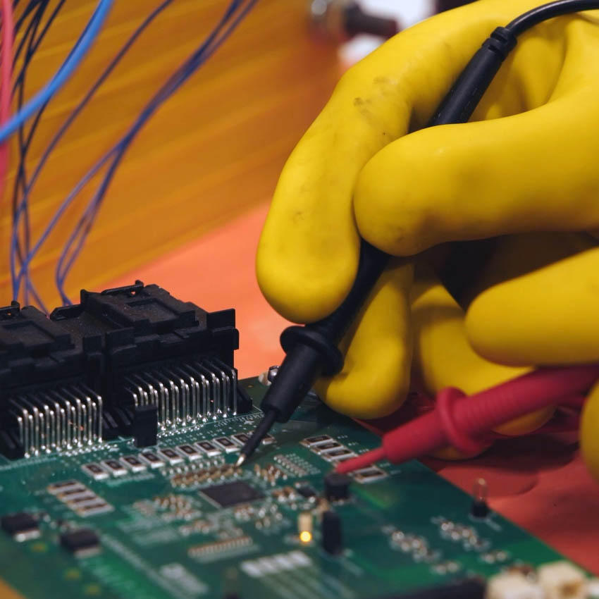 Yellow-gloved hand uses multimeter probe on circuit board.