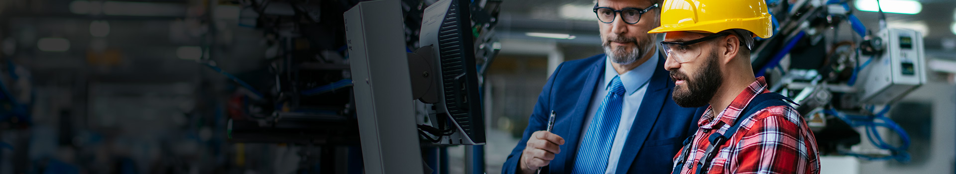 Two engineers in a manufacturing facility, one in a suit holding a clipboard and the other in a hard hat and safety gear operating a machine through a computer terminal.