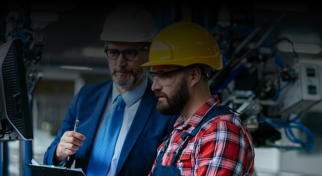 Two engineers in a manufacturing facility, one in a suit holding a clipboard and the other in a hard hat and safety gear operating a machine through a computer terminal.