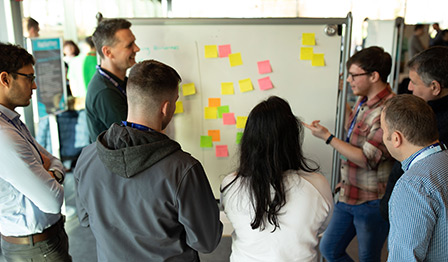 A group of colleagues standing in a circle around a whiteboard covered with colorful sticky notes, engaging in a collaborative brainstorming session.