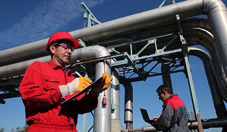 Two industrial workers wearing protective gear conducting inspections at an outdoor facility with large metal pipes and infrastructure.