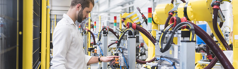 Man in a factory inspecting industrial robots;  he's checking wiring and controls on yellow robotic arms.