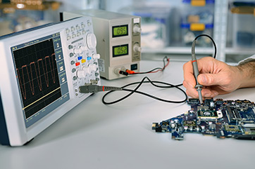 Technician using an oscilloscope and probe to test a circuit board in a lab environment, with a power supply in the background displaying voltage readings.