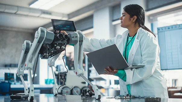 A female robotics engineer in a lab coat using a laptop while examining a four-legged robot dog.