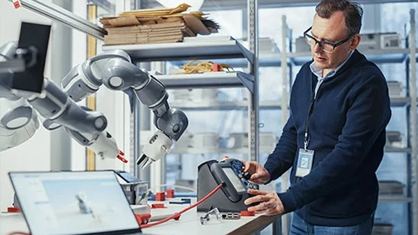 Man with glasses operates a robotic arm in a lab with laptop and tools.