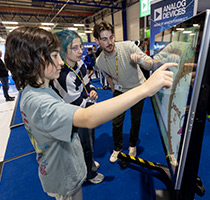 Two children using an interactive touchscreen STEAM kiosk at an Analog Devices event, guided by a staff member.
