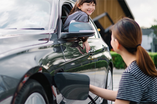 a mother plugging an electrical charger into her car while laughing with her daughter in the car window