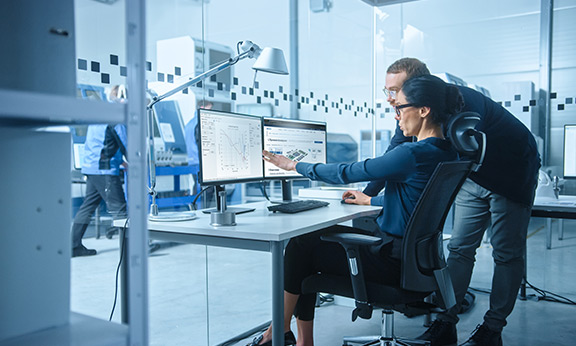 Two engineers collaborate on data displayed on dual monitors.