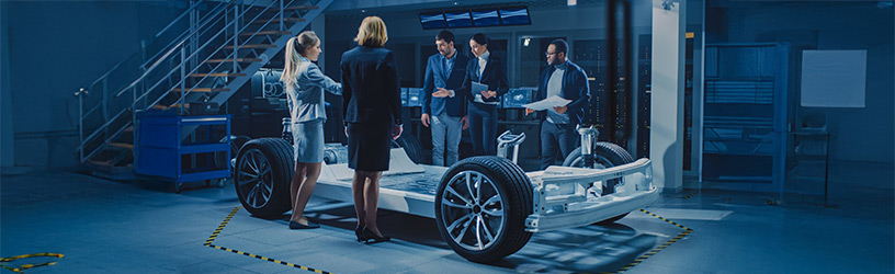 Team of automotive engineers reviewing an electric vehicle chassis and battery platform in a high-tech development facility
