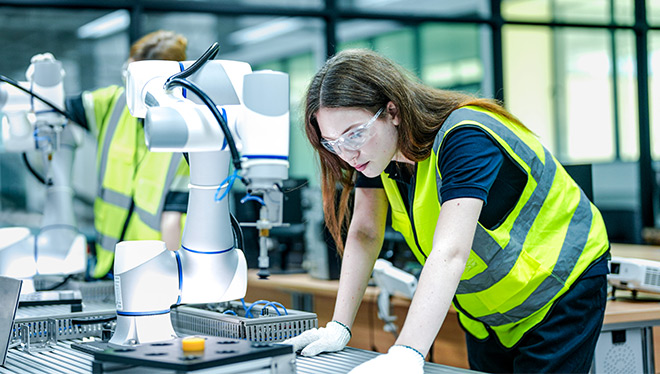 Woman in high-vis vest and glasses working with robotic arms.