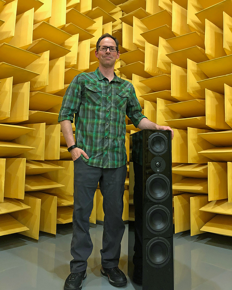 A man standing in an anechoic chamber next to a floor-standing speaker