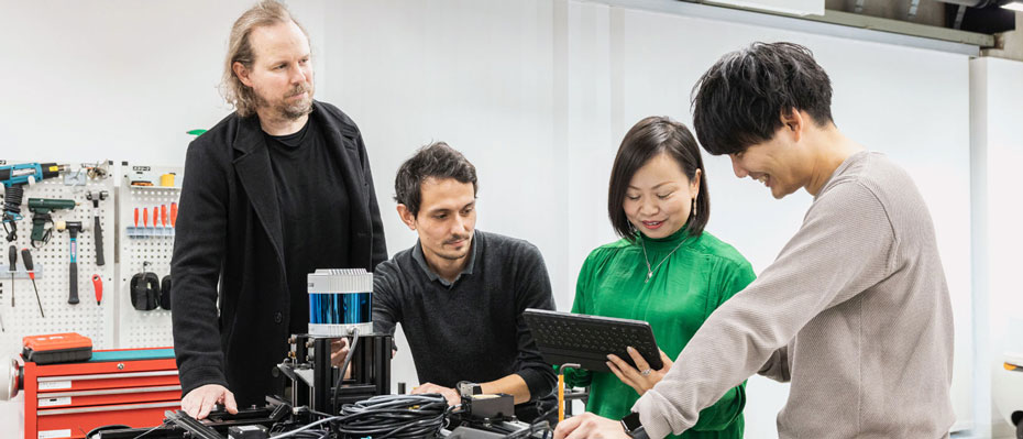 Four people collaborating in a workshop with tools. One holds a tablet while others discuss.