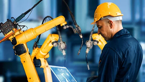 Man in hard hat monitors yellow industrial robots with a tablet.