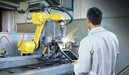 Engineer using a tablet to control or monitor a robotic arm performing automated welding in a manufacturing environment.