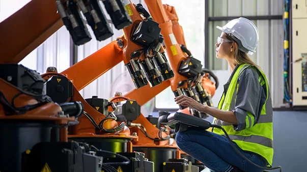 Woman in safety gear operates an industrial robot arm with a handheld controller in a factory.