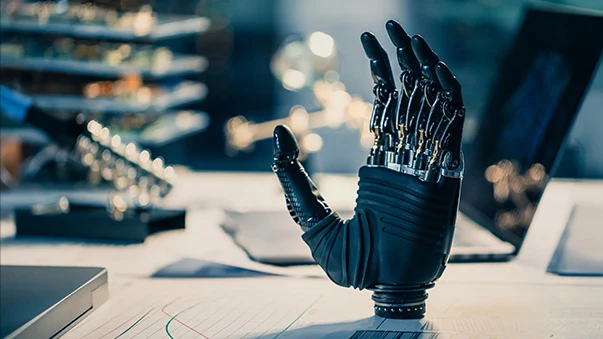 Black robotic hand with exposed parts on a desk with papers in a lab.