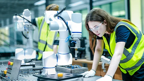Woman in safety vest and glasses inspecting a robotic arm.