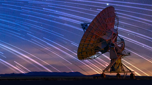 Large satellite dish with star trails in the night sky behind it.