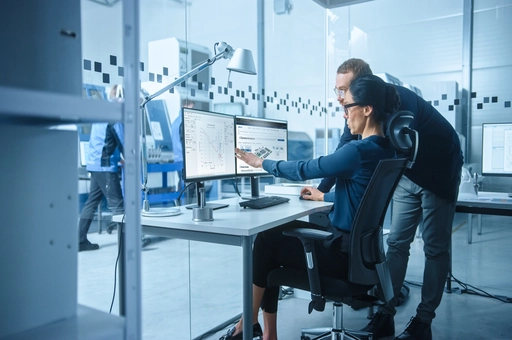 Two engineers collaborate on data displayed on dual monitors.