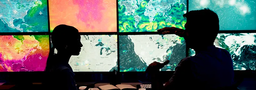 Two people analyze weather data on a wall of monitors.