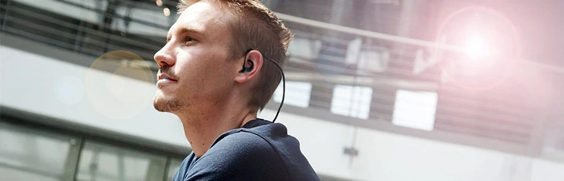 Man wearing black neckband earphones, looking thoughtfully upward in a modern building.