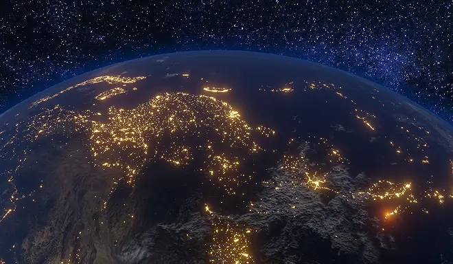 View of Earth from space at night, showing illuminated city lights across Europe and surrounding regions, with a star-filled sky in the background.