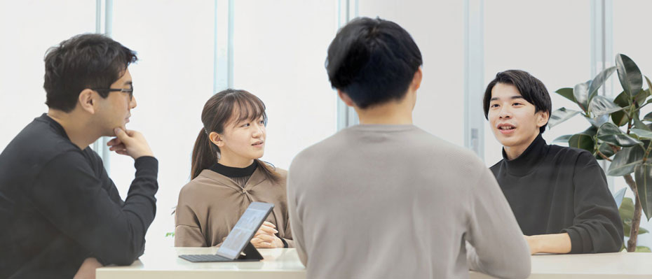 Four people in a meeting around a table with a tablet, potted plant, and natural light from large windows.