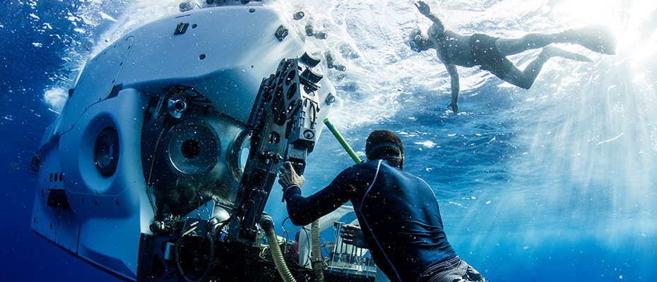 Image shows underwater ocean scene with two scientists manuvering around in the water with snorklers and fins; along with a large data-gathering piece of equipment.