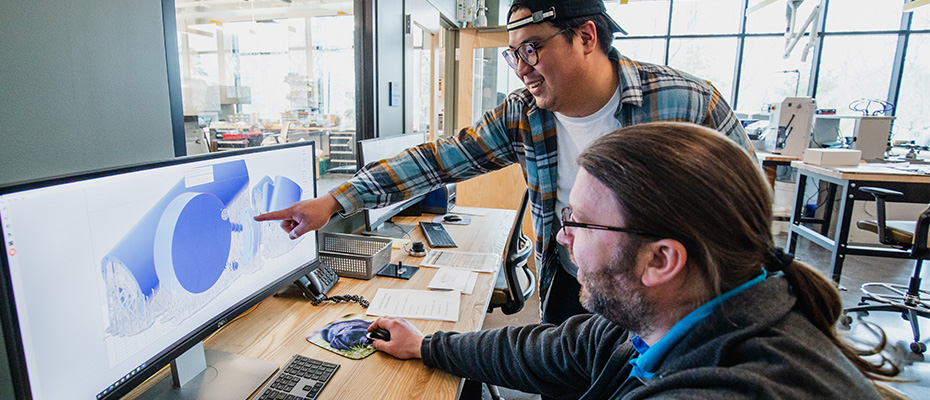 Image shows two male workers in an office laboratory setting. One person is pointing at a computer monitor and both are discussing the data in the monitor.