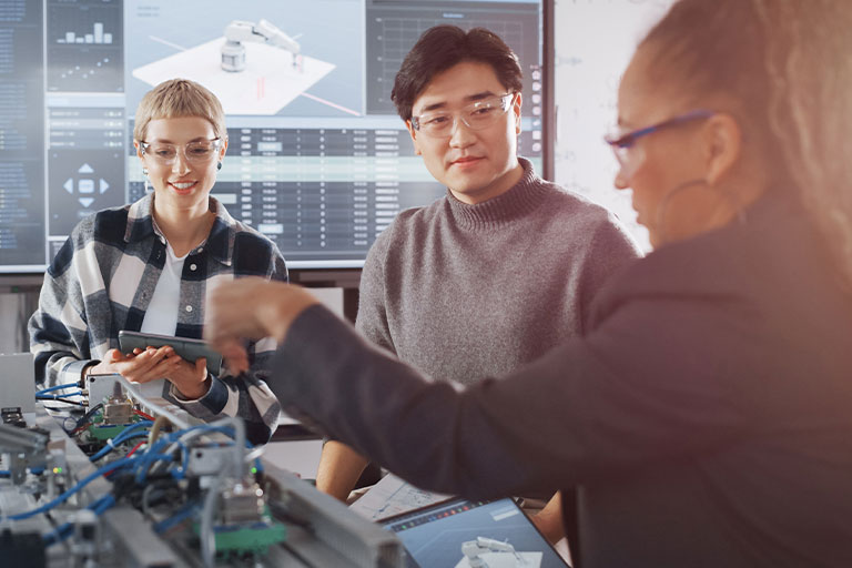 Three people collaborate in an office with machinery and technical data screens.