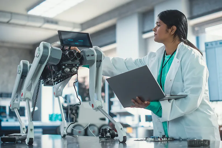 A female robotics engineer in a lab coat using a laptop while examining a four-legged robot dog.
