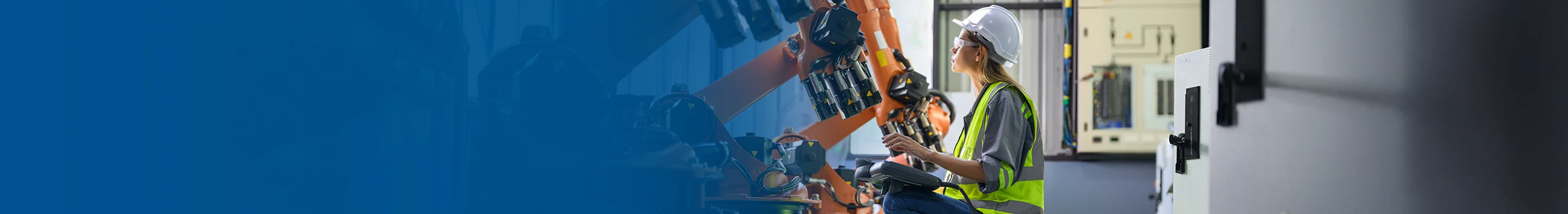 Engineer in hard hat and safety vest kneels, operating a control panel near several large orange industrial robots in a factory.