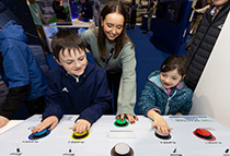 Children playing with colorful interactive buttons at an arcade corner, supervised by a smiling woman.