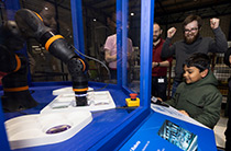 Child operating a robotic arm at an Analog Devices exhibit while onlookers cheer in excitement.