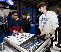Two children interacting with a sound synthesizer exhibit at an Analog Devices booth, guided by a staff member.