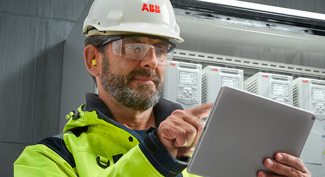 Industrial worker in safety gear using a tablet in front of ABB electrical equipment.
