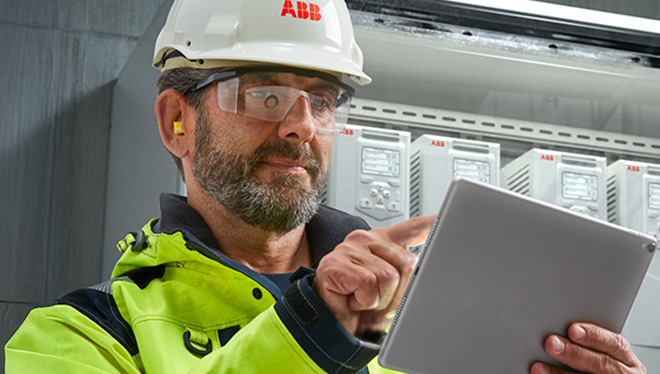 Industrial worker in safety gear using a tablet in front of ABB electrical equipment.