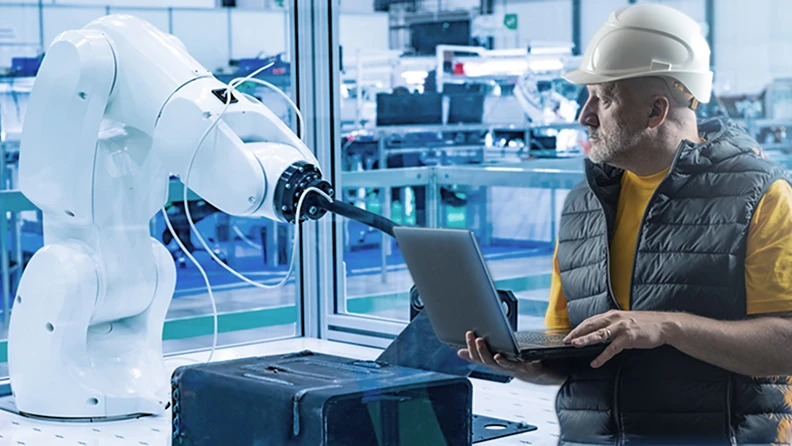 Engineer with laptop observing robotic arm in a factory.