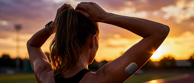 Sweaty person with CGM on arm, tying hair, at sunset.
