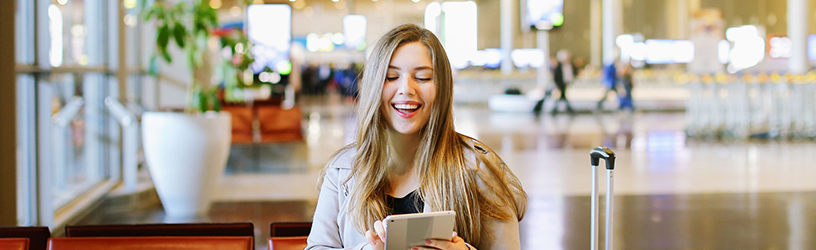 Smiling woman sitting in an airport terminal, using a tablet with her luggage beside her, enjoying her time while waiting for her flight.