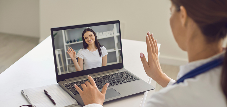 Female nurse looking at laptop during a remote meeting with a female patient. Both waving hello.