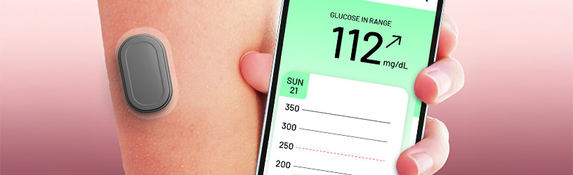 Close-up of glucose sensor on arm with smartphone displaying 112 mg/dL glucose reading