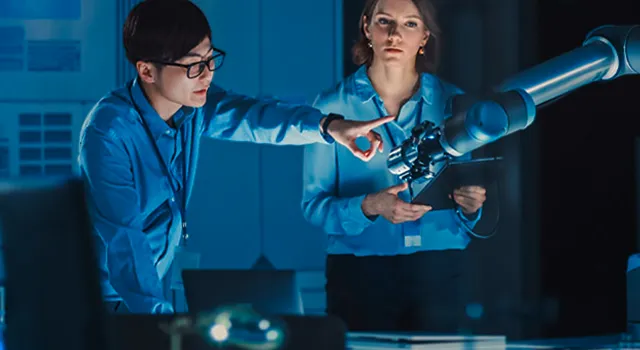 Two engineers collaborate with a robotic arm in a blue-lit lab.