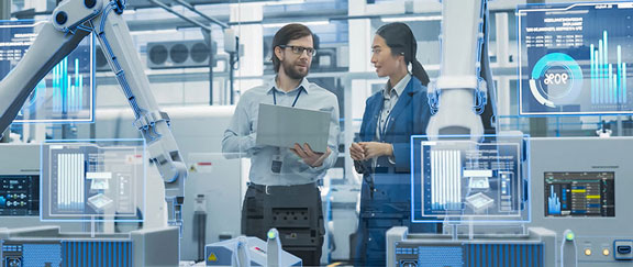 Engineers in futuristic factory discuss with laptop, surrounded by robotic arms and holographic displays.