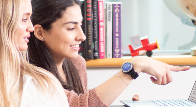 Image shows two people sitting on a couch looking at a computer laptop monitor while one one points at the screen.