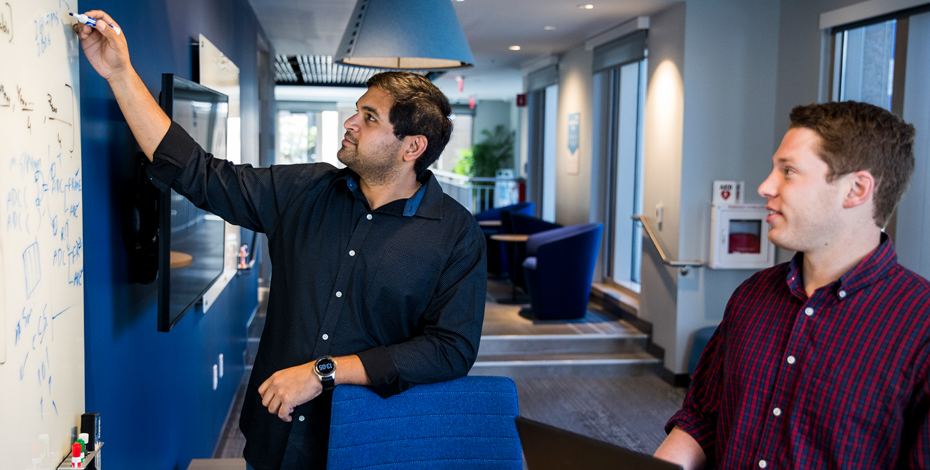 Image shows two men collaborating in an office-setting. They are discussing and writing technical data on a whiteboard located on a wall infront of them.