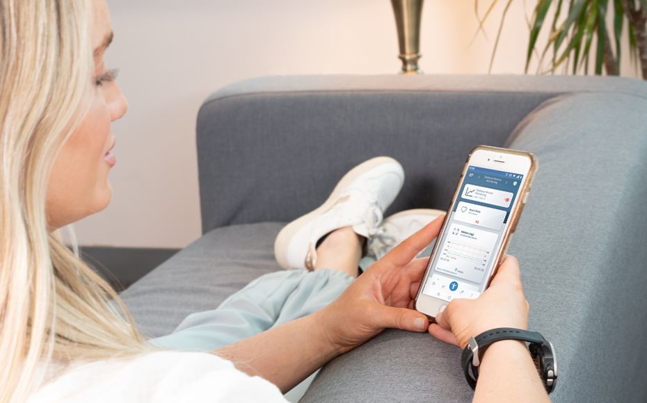 Image shows a woman sitting on a couch while looking her cel phone. The Cel Phone screen shows data from the Vitruvian Shield Monitoring Application.