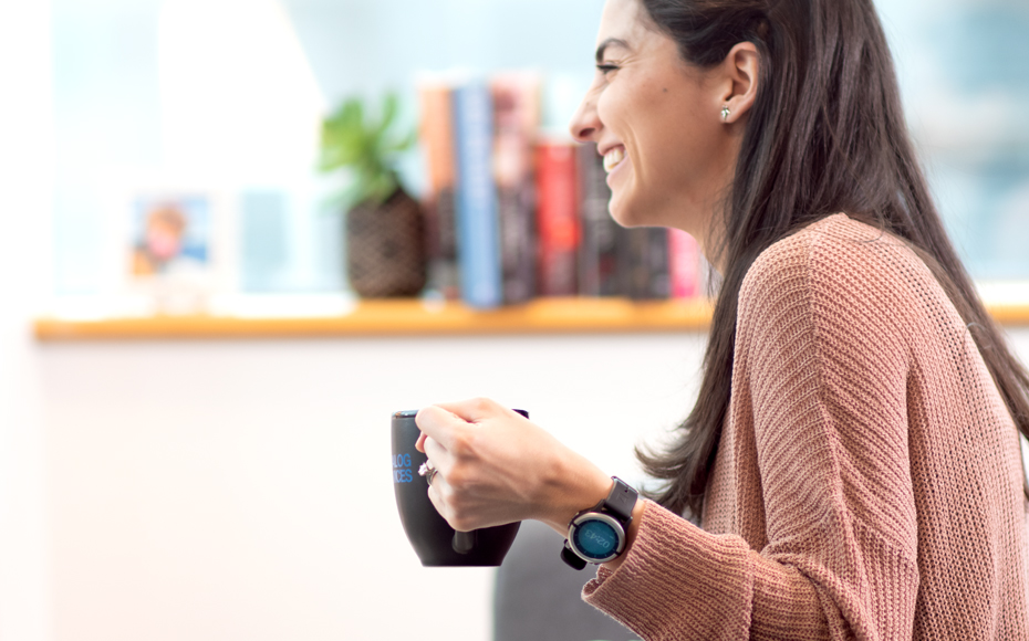 Image shows a woman smiling and enjoying a cup of coffee. She is wearing a Vitruvian Shield monitoring watch.